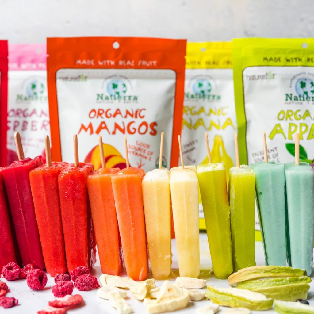 A rainbow spectrum of popsicles sit before an assortment of Natierra freeze dried berry bags.